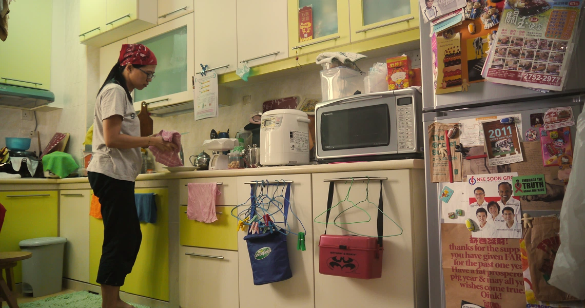 A woman stands in a cramped kitchen from the film "03-Flats", surrounded by appliances and colorful decor.