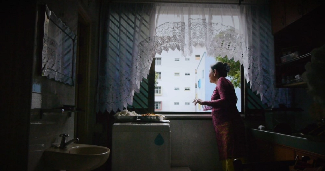 A dimly lit room in "03-Flats" film, showing a figure in red by a window with lace curtains and urban view outside.