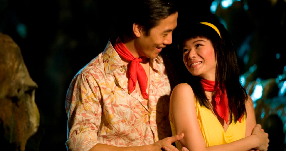 A film still from "12 Lotus" showing two characters embracing and smiling, with one wearing a floral shirt and red scarf.