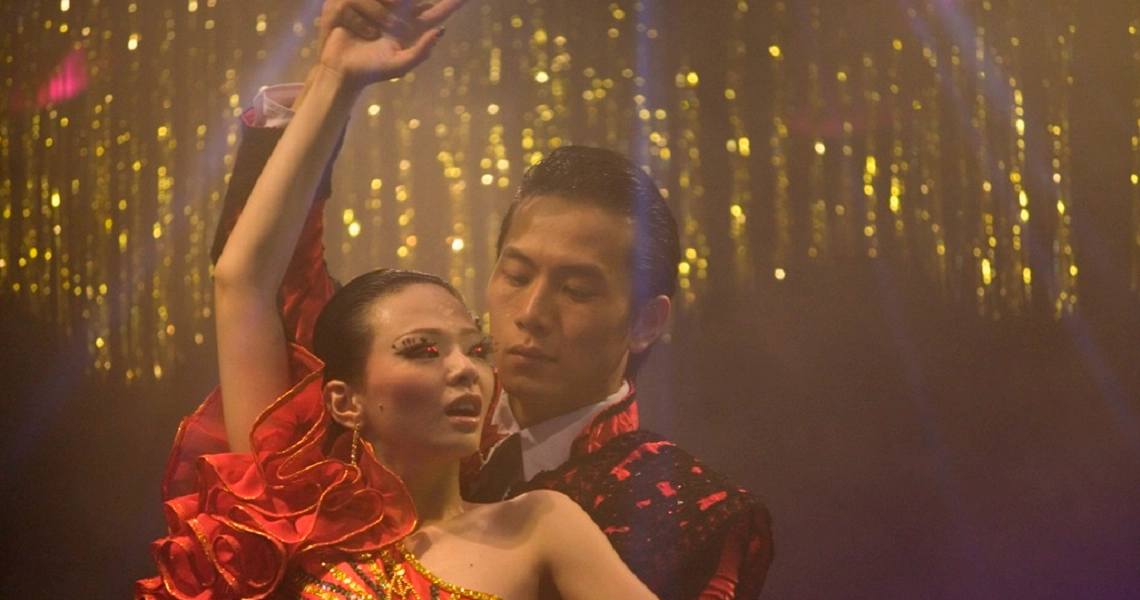 A couple performs a passionate dance in "12 Lotus," with the woman in a vibrant red dress against a glittering golden backdrop.