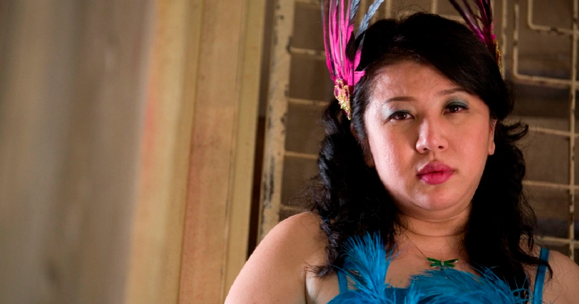 A woman in a blue dress with a pink feather headpiece from the film "12 Lotus" looks pensively to the side.