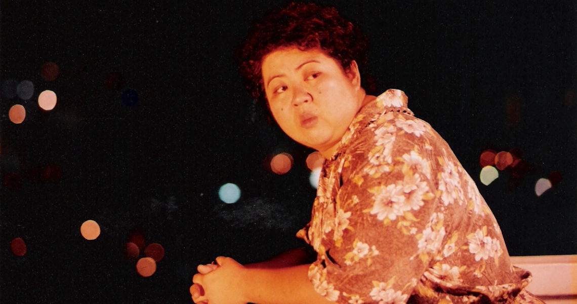 A film still from "12 Storeys" showing a person in a floral shirt against a dark background with blurred lights.