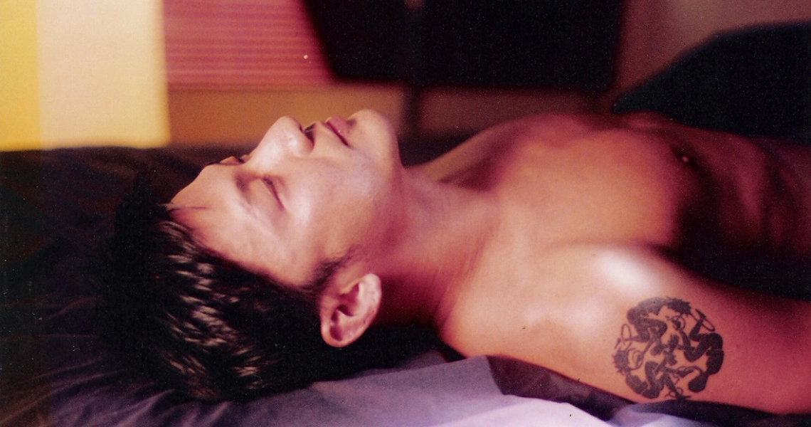 A film still from "12 Storeys" showing a shirtless man with a tattoo lying down, eyes closed, in warm lighting.