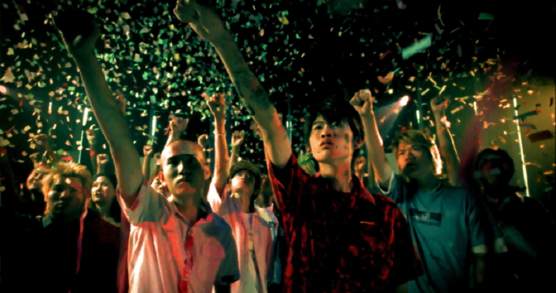 A crowd of young people with raised arms celebrate amidst falling confetti in a vibrant scene from the film "15".