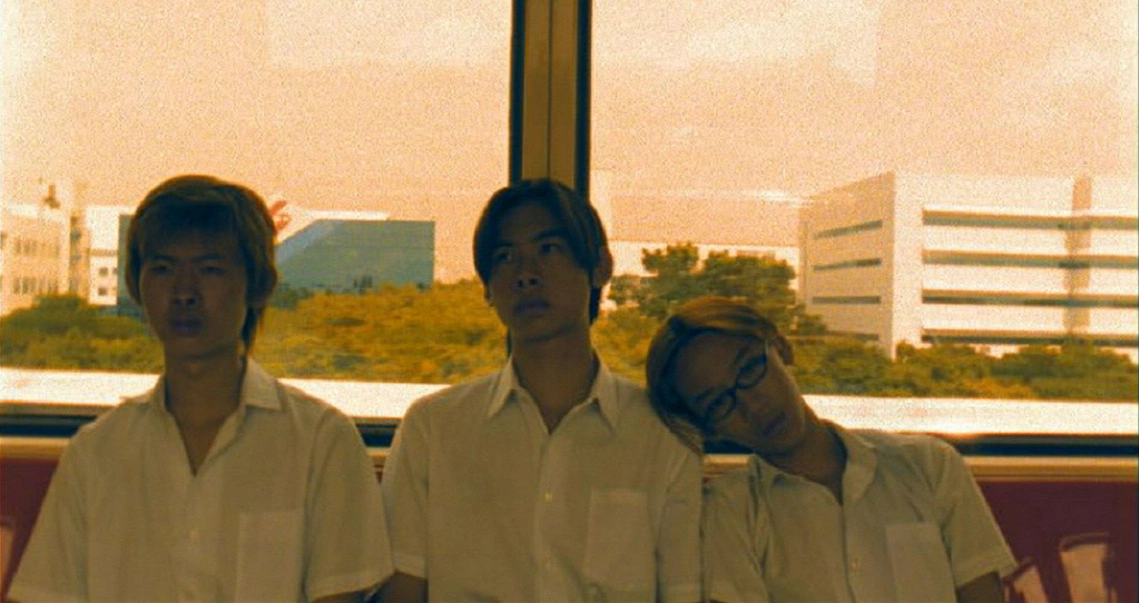 Film still from "15" showing three young men in white shirts sitting together, with urban buildings visible in the background.