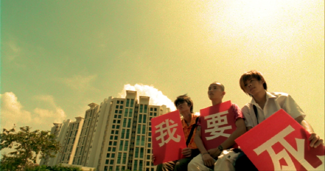 Film still from "15" shows three young men holding red signs with Chinese characters against a backdrop of high-rise buildings.