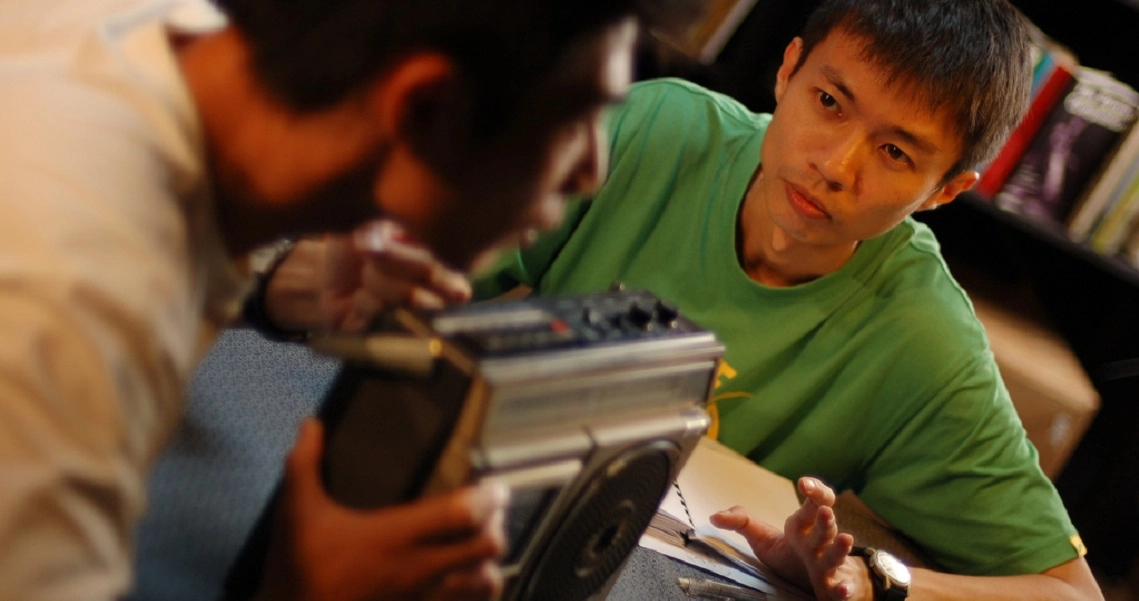 A film still from "18 Grams of Love" showing a young man in a green shirt intently working with electronic devices.