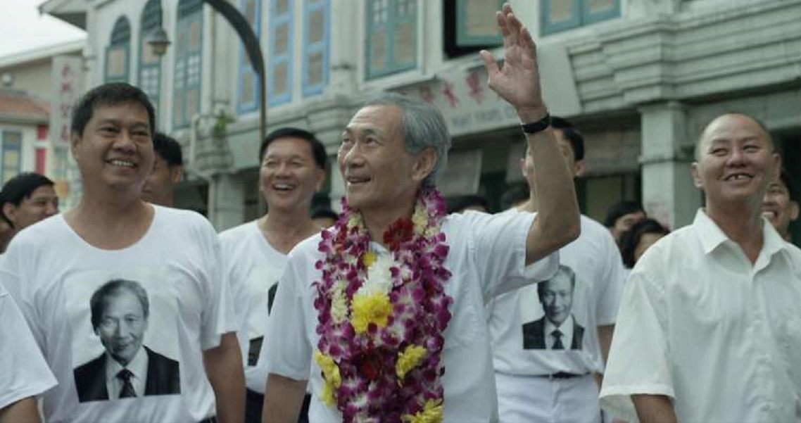 A scene from the film "1965" showing a smiling man with a floral garland surrounded by supporters wearing t-shirts with a portrait.