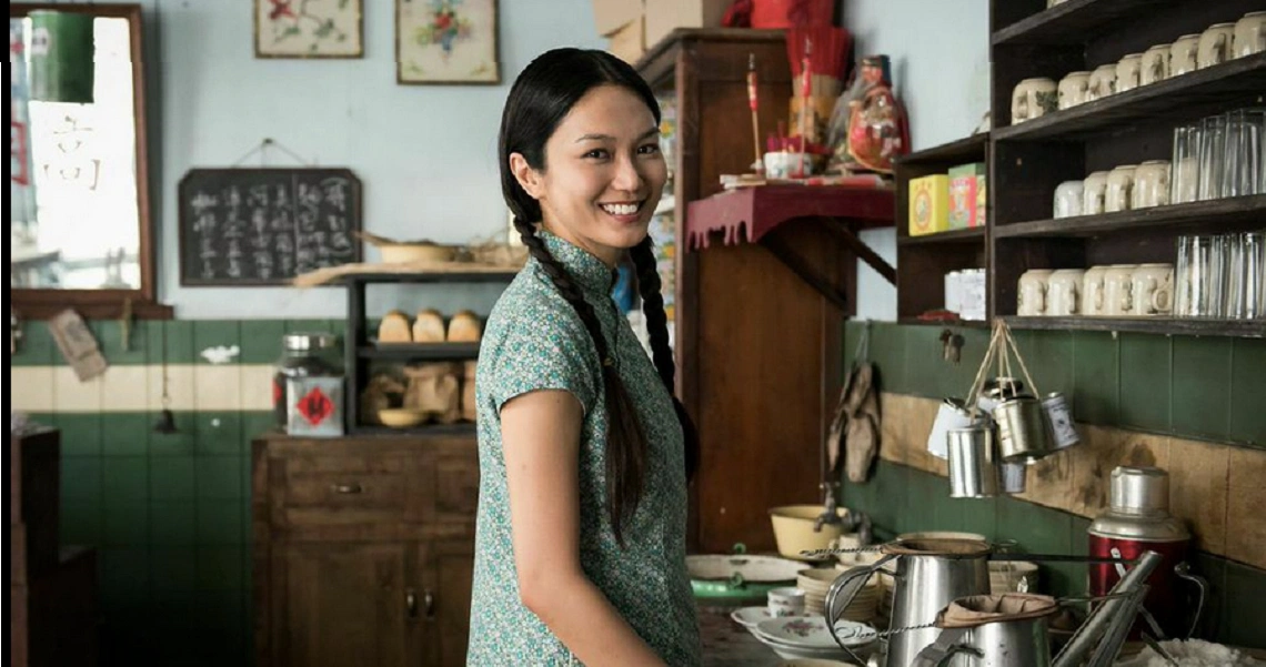 A scene from the film "1965" shows a woman in traditional attire smiling in a vintage Singaporean kitchen filled with period details.