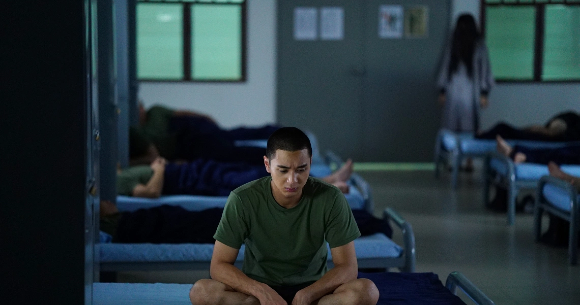 A tense scene from the film "2359: The Haunting Hour" shows a man sitting on a bed in a dimly lit dormitory with other occupied beds visible.