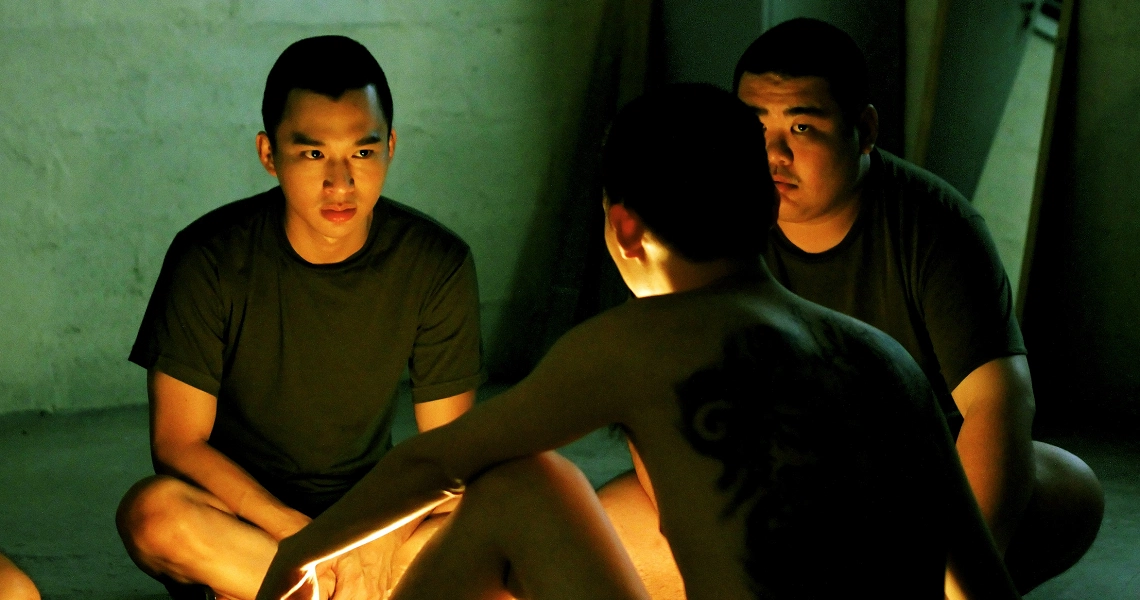 A tense scene from the film 2359 shows three young men in dark t-shirts huddled around a glowing light source in a dimly lit room.