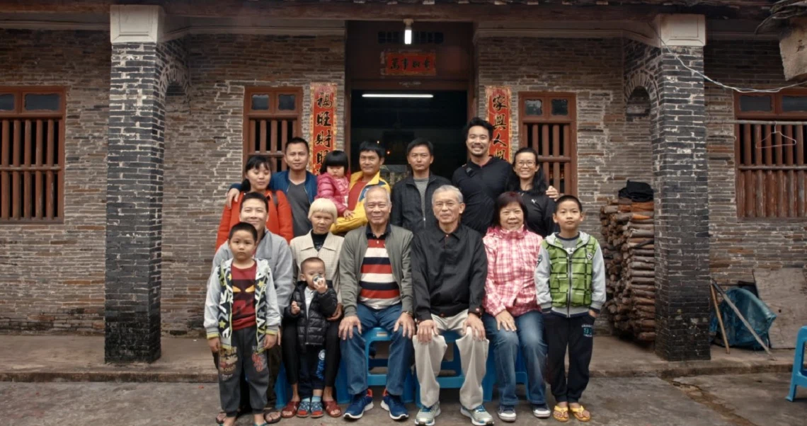 Film still from "667" showing a large family group posed in front of a traditional Chinese brick house with wooden doors and tiled roof.