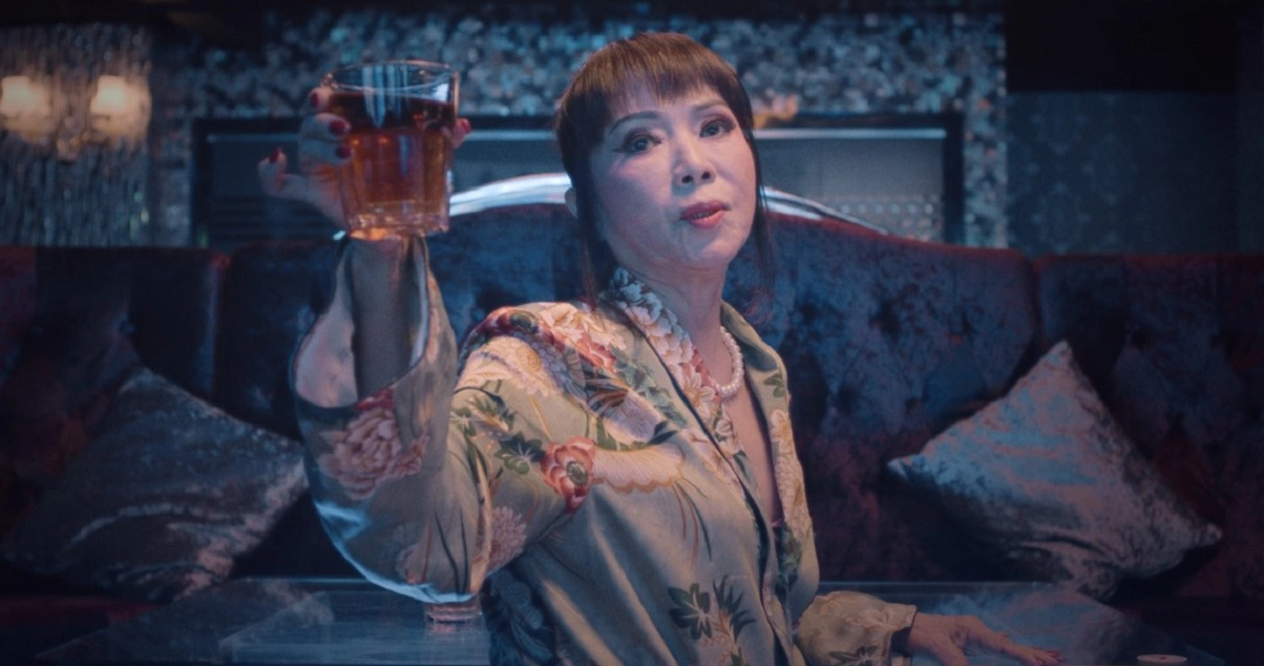 A woman in a floral robe holds up a glass of wine, looking contemplative in a dimly lit room. Film still from "667".