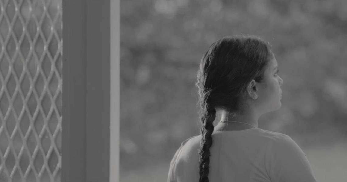 A young girl with a braided hairstyle stands near a window in a scene from the film "7 Letters".