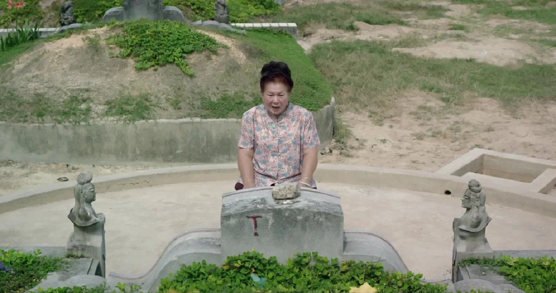 A scene from the film "7 Letters" showing a man in a patterned shirt sitting at a concrete table with small statues in a garden setting.