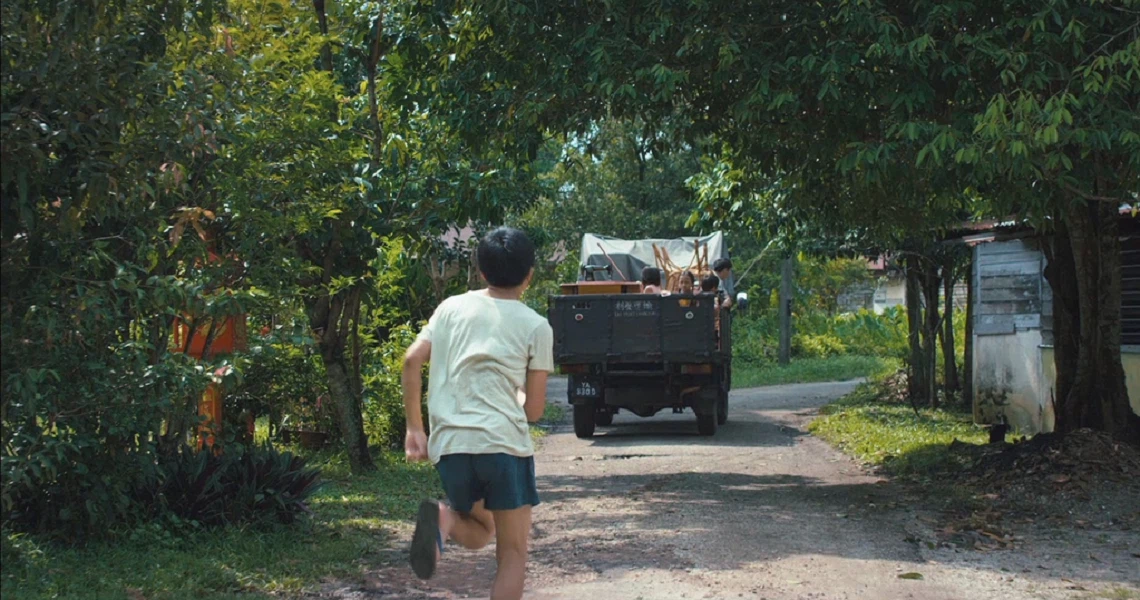 A scene from the film "7 Letters" showing a person walking behind a truck on a rural path surrounded by lush greenery.