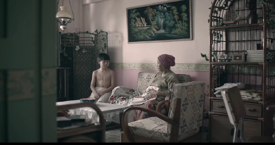 A scene from the film "7 Letters" showing two people seated in a vintage-style room with ornate furniture and a decorative screen.