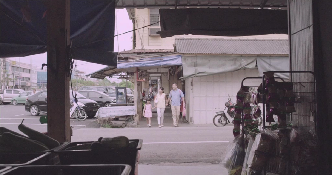 A street scene from the film "7 Letters" showing people standing near buildings and parked vehicles in a vintage-style setting.