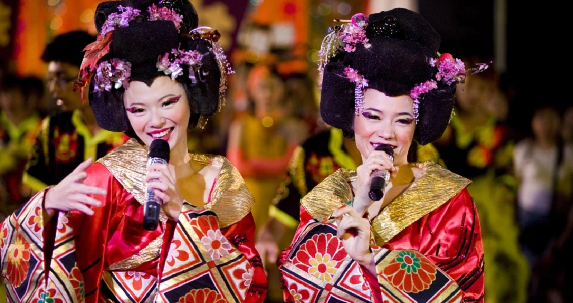 Two performers in colorful geisha-inspired costumes sing on stage in a scene from the Singaporean film 881.