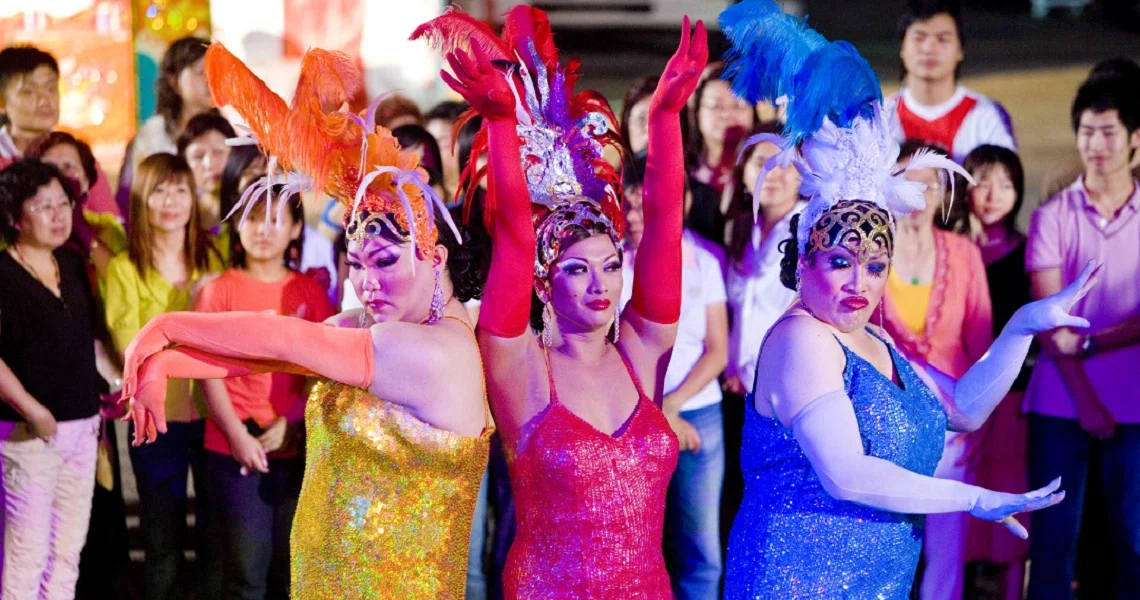 Colorful scene from the film 881 showing three Getai performers in vibrant sequined dresses dancing with raised arms, surrounded by audience members.