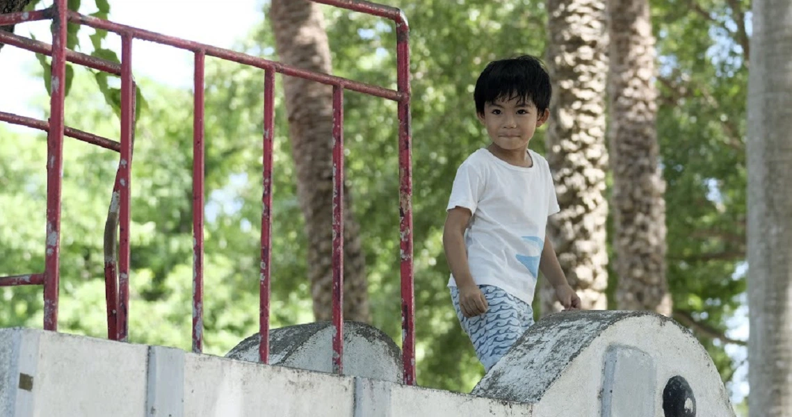 A young boy in a white shirt stands on a concrete structure in a scene from the film "A Family Tour".