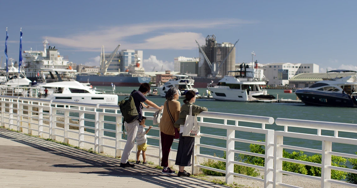 Three people stand on a pier overlooking a marina with yachts and ships in the film "A Family Tour".