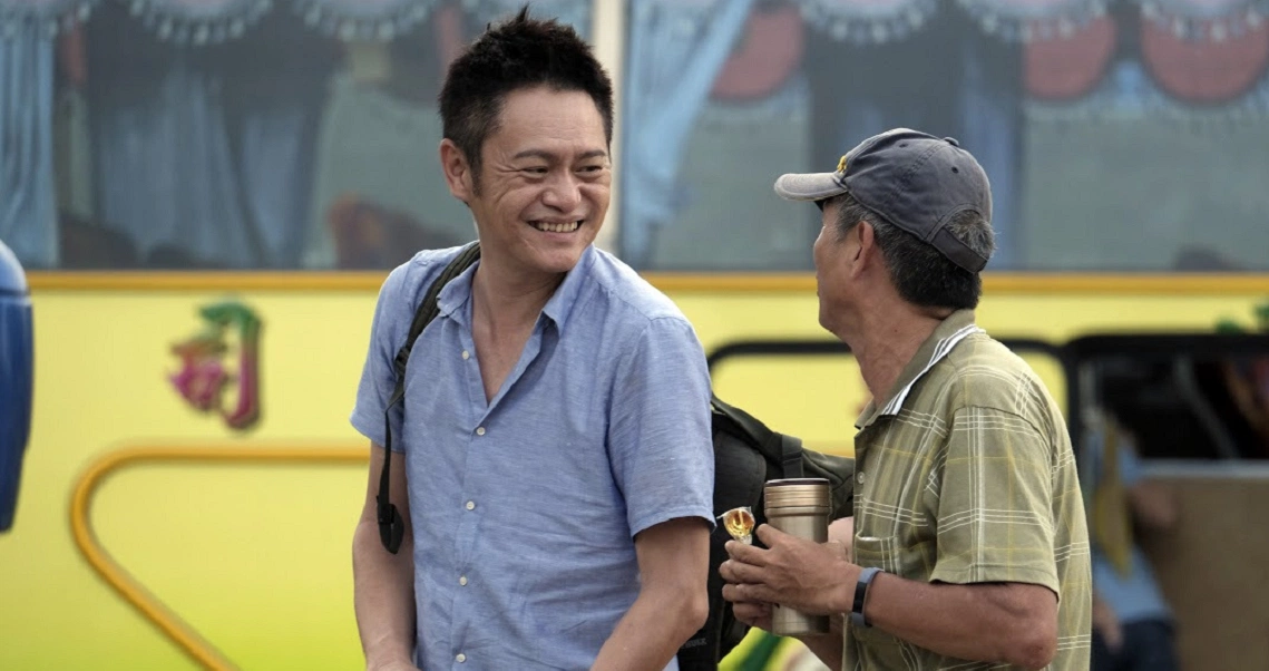 A scene from the film "A Family Tour" showing two men in casual attire conversing outdoors near a yellow vehicle.