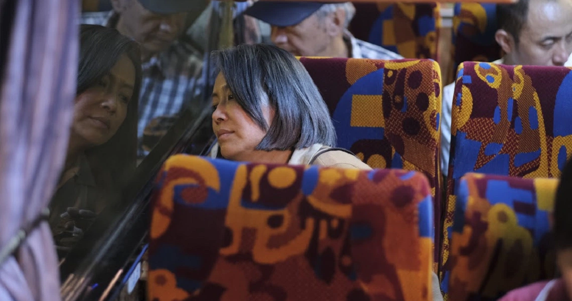 A woman with short hair sits on a colorful bus seat in a scene from the film "A Family Tour".