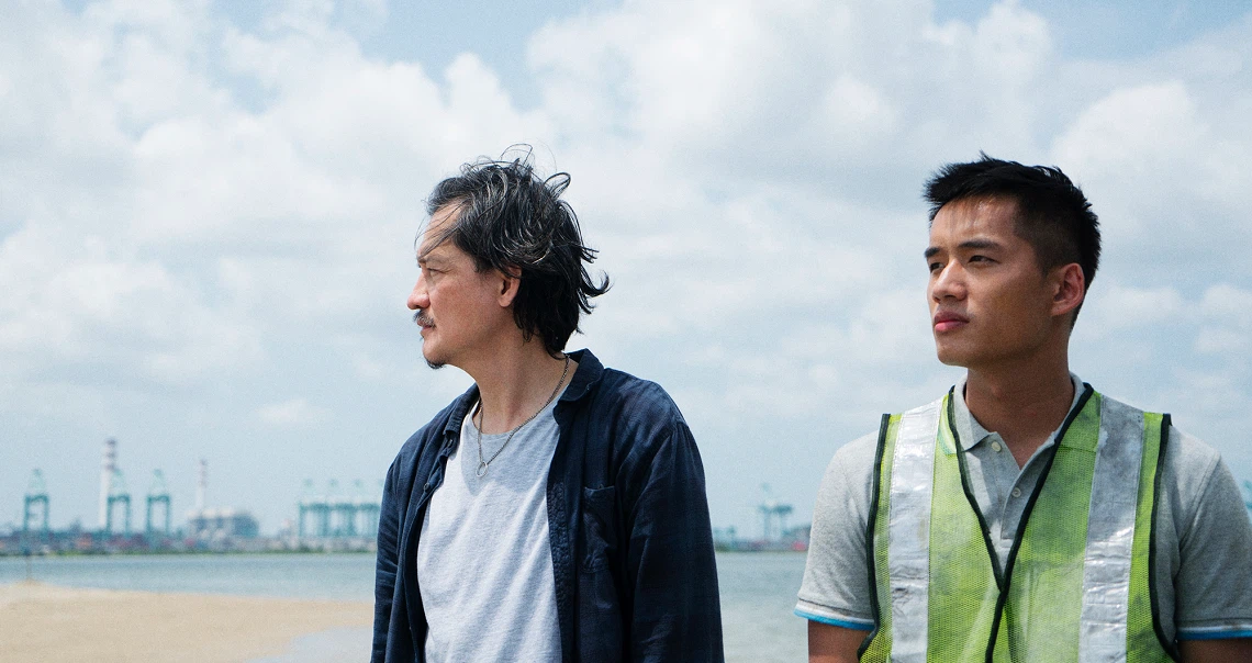 Film still from "A Land Imagined" showing two men standing on a beach with industrial cranes visible in the background.