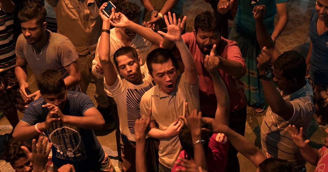 A Land Imagined film still shows a lively crowd of young men dancing and raising their hands in a dimly lit, energetic scene.