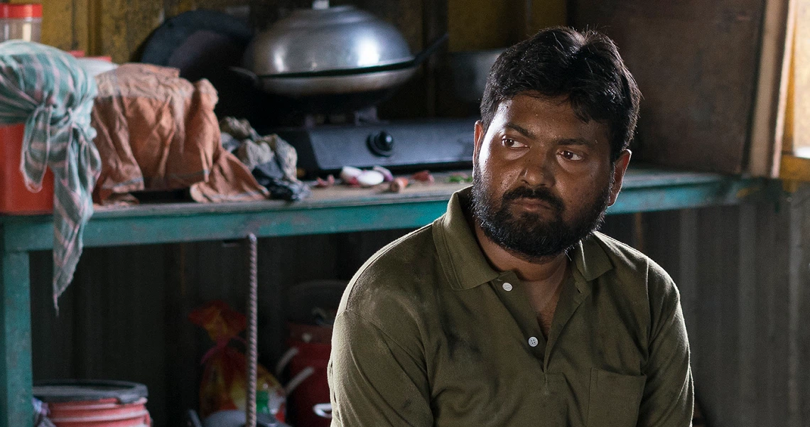 A film still from "A Land Imagined" showing a bearded man in a green shirt sitting in a cluttered, dimly lit room.