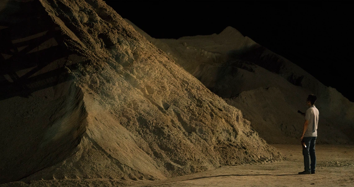 A still from "A Land Imagined" shows a person standing before massive sand piles at night, capturing the film's industrial landscape.