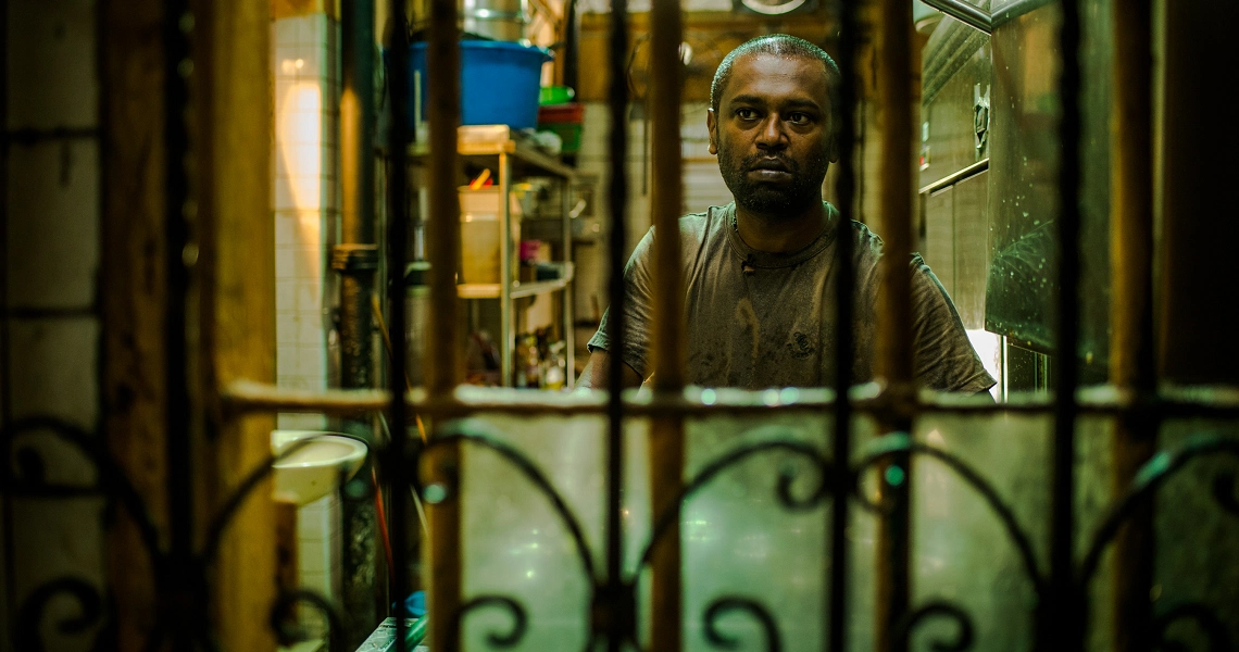 A film still from "A Yellow Bird" showing a man peering through an ornate metal gate in a dimly lit, narrow alleyway.