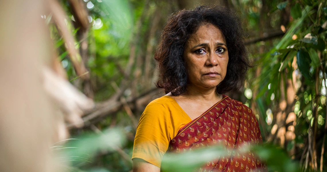 A film still from "A Yellow Bird" shows a woman in a yellow and red sari looking concerned amidst lush greenery.