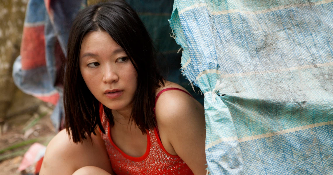 A film still from "A Yellow Bird" shows a woman in a red top sitting next to a tattered blue tarp, conveying a sense of hardship.