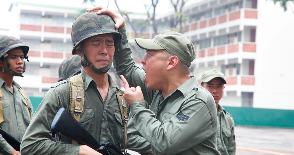 A scene from "Ah Boys to Men" shows military recruits in uniform, with one soldier adjusting another's helmet during training.