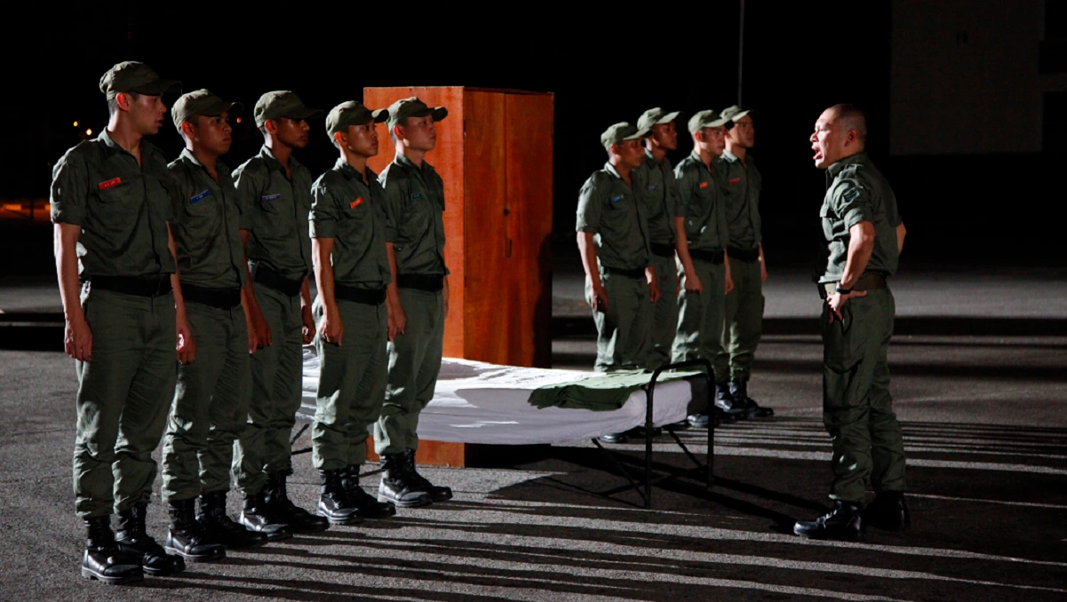 A scene from the film "Ah Boys to Men" showing military recruits standing at attention during a night-time assembly.