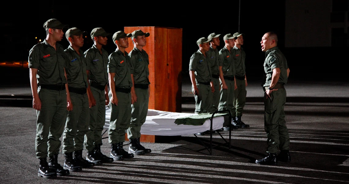 A scene from the film "Ah Boys to Men" showing military recruits standing at attention during a night-time assembly.