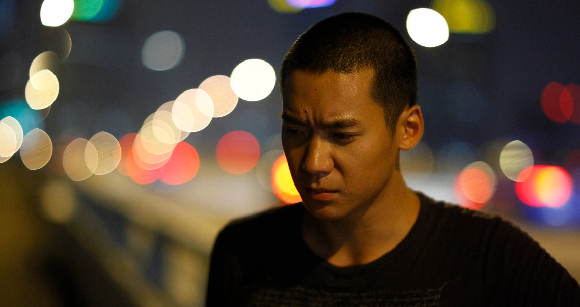 A young man from the film "Ah Boys to Men" looks pensive at night, with blurred city lights in the background.