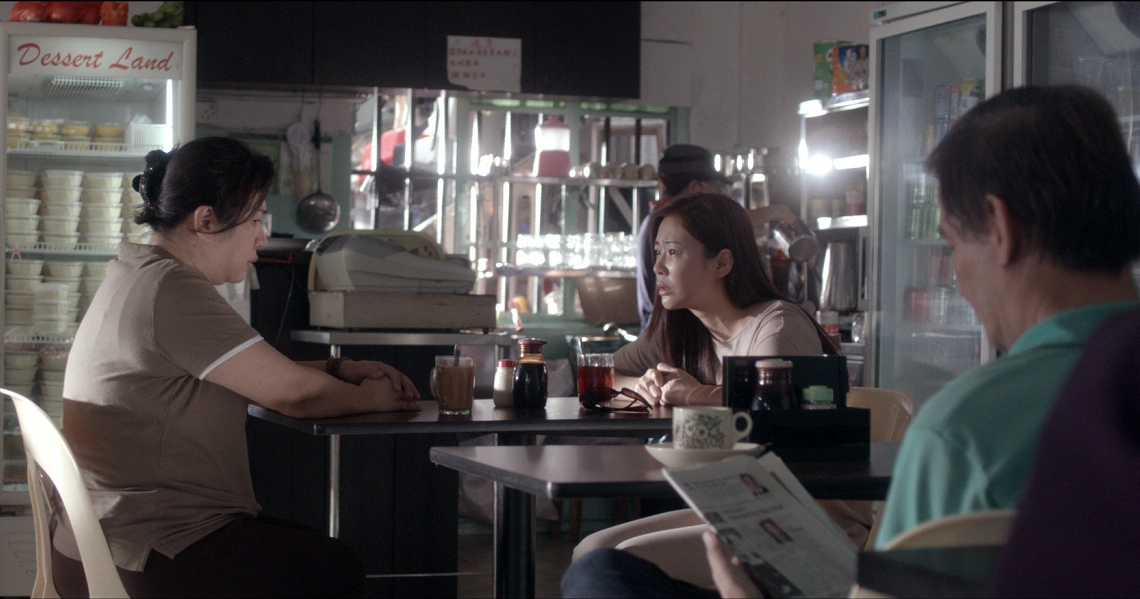 A tense scene from "Bring Back The Dead" shows two women in conversation at a cafe table, with a man reading nearby.