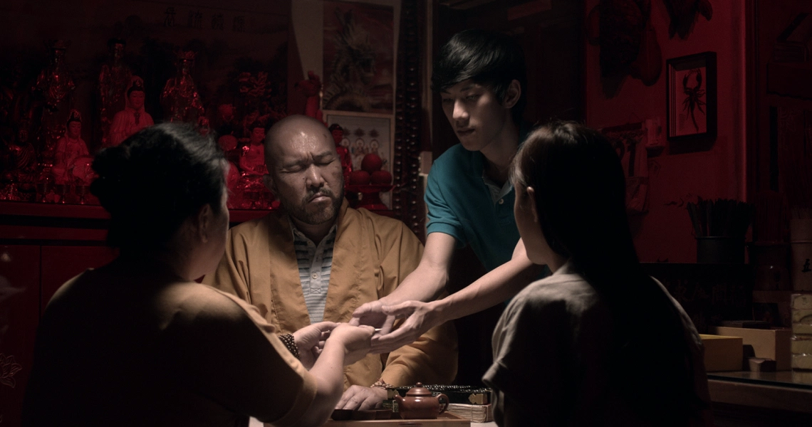 A tense scene from "Bring Back The Dead" shows four people in a dimly lit room with religious statues, engaged in a spiritual ritual.