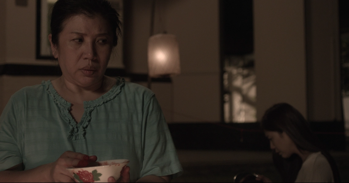 A scene from "Bring Back The Dead" showing a woman in a green top holding a floral bowl, with a dimly lit interior in the background.