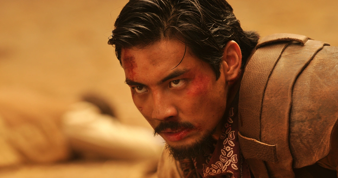 A close-up of a rugged, bearded man with intense eyes in the film Buffalo Boys. He appears to be in a dusty, desert-like setting.