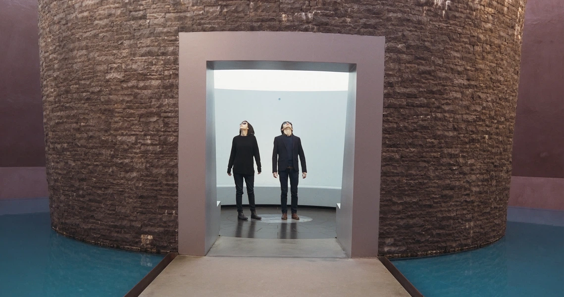 A film still from Cannonball showing two figures standing in a doorway of a circular stone structure surrounded by blue water.