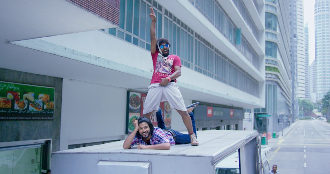 A scene from Chennai2Singapore film showing characters posing playfully on a ledge in front of a modern building with skyscrapers in the background.