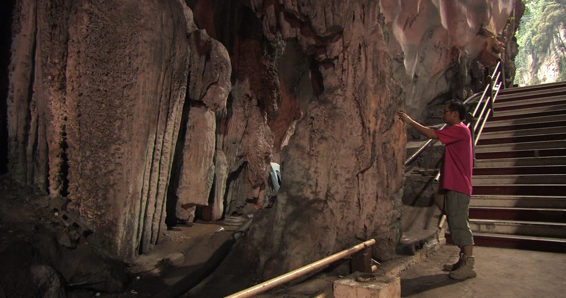 A person in red shirt gestures towards cave formations in a scene from the film "Dreams From The Third World".