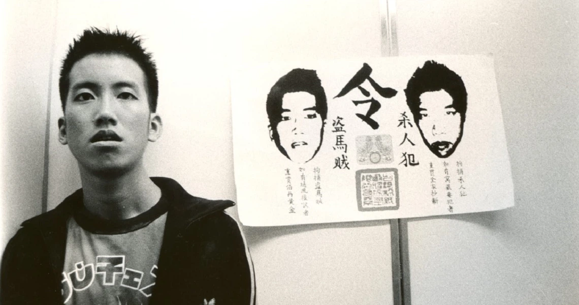 A black and white film still from "Eating Air" showing a young man next to a poster with illustrated faces.