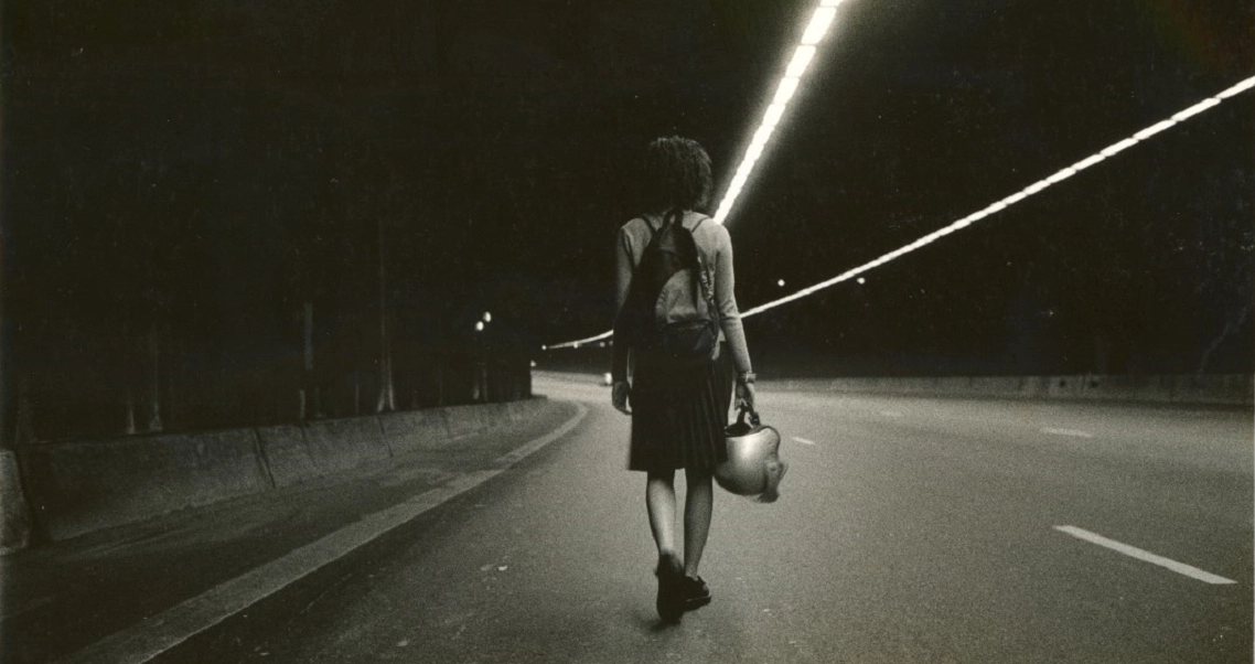 A film still from "Eating Air" showing a person walking alone on an empty road at night, illuminated by streetlights.