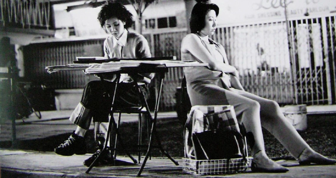 A black and white film still from "Eating Air" showing two young people sitting on chairs outdoors, capturing a moment of urban youth culture.