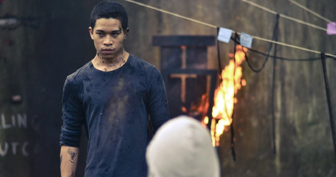 A tense scene from the film Faeryville shows a character in a dirty shirt standing near flames, creating a dramatic atmosphere.